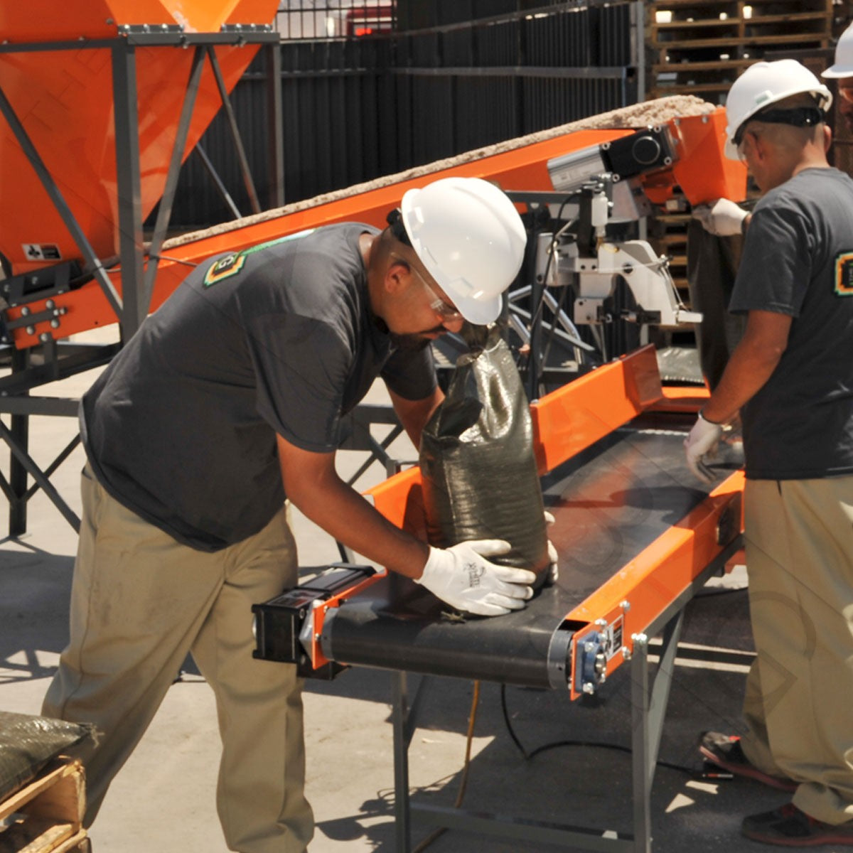 Three person crew in white hard hats operating the sandbag sewing table on a bagging yard in full sun
