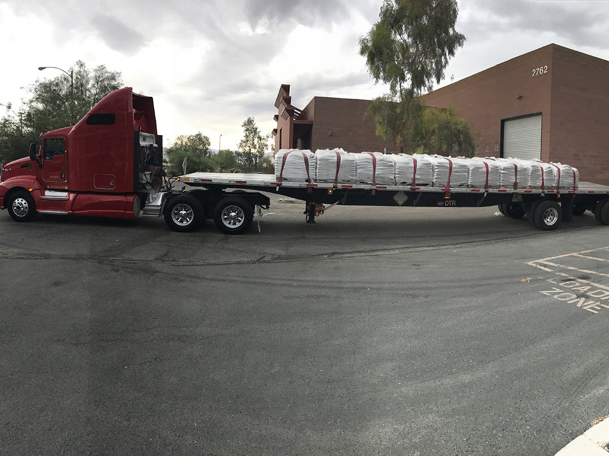A flatbed semi-truck full of 18 pallets of filled sandbags with red straps leaving the bagging yard