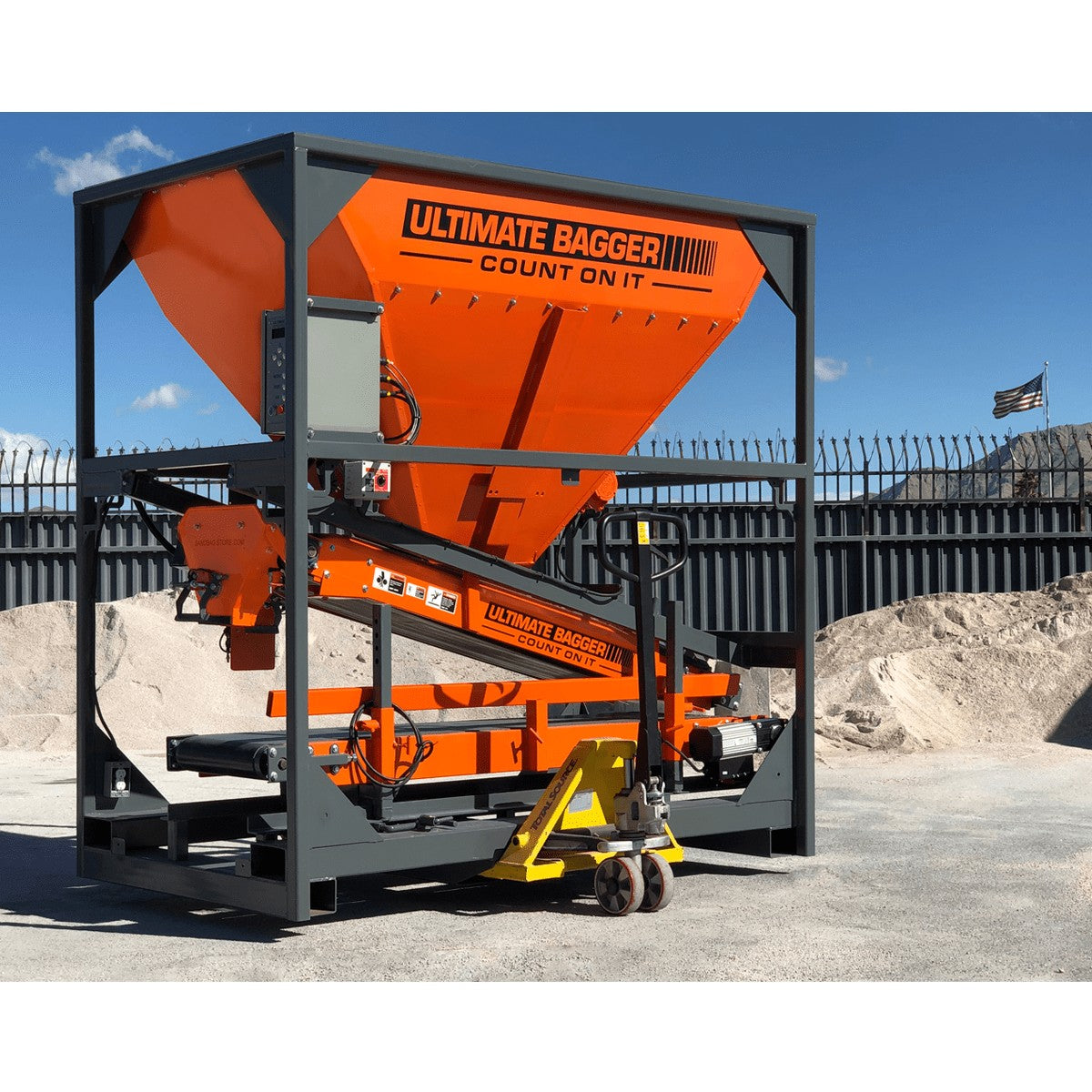 Image of the Ultimate Bagger fully packed with lower bag closing table stowed and bag filling spout retracted being moved with a simple pallet jack on a bagging yard with piles of sand and the american flag in the background